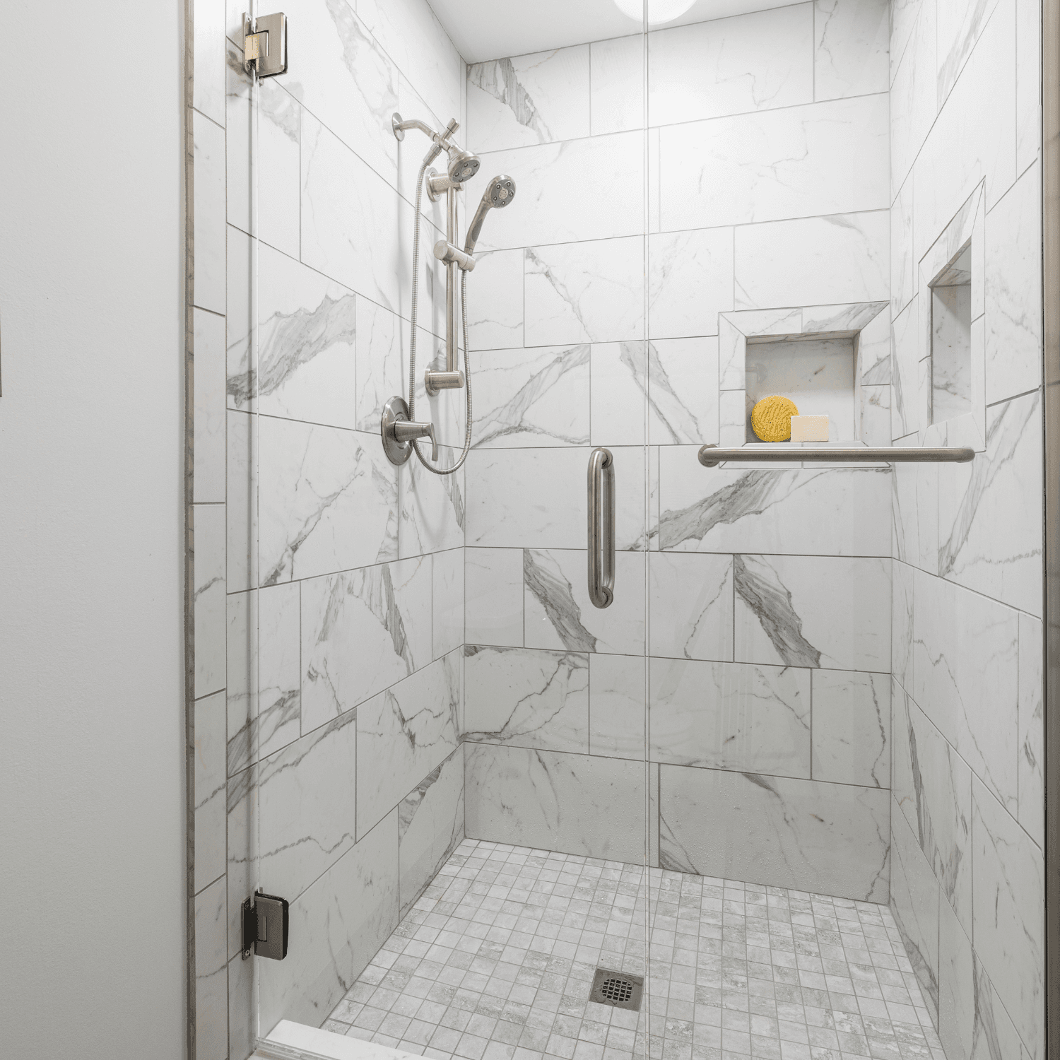 Marble tile walk-in shower with glass enclosure, dual showerheads on brushed nickel fixtures, built-in shelf holding yellow washcloth and soap, and floor drain